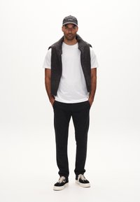 Graue Strickweste über einem weißen T-Shirt, kombiniert mit schwarzen Sweatpants und schwarzen Sneakern mit weißen Sohlen. Die Mütze ist dunkel und trägt ein Logo.