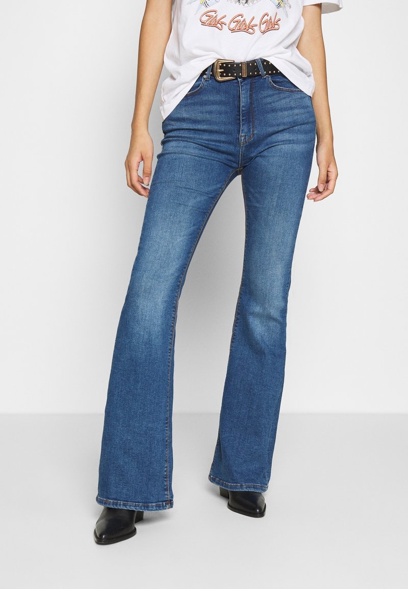Femme portant un jean flare taille haute bleu, un t-shirt blanc à motif rentré, une ceinture cloutée noire et des bottines noires.