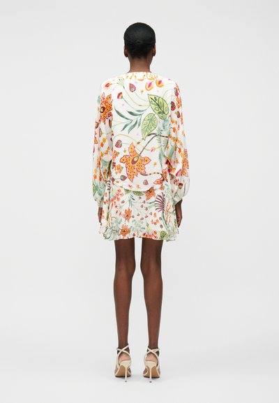 Γυναίκα με σκούρο δέρμα και κοντά μαλλιά φοράει λευκό φόρεμα με μακριά μανίκια και φλοράλ σχέδιο, καθώς και μπεζ ψηλοτάκουνα σανδάλια, στέκεται με την πλάτη σε απλό φόντο.