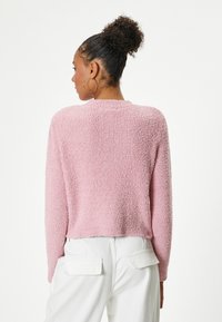 Pull en fausse fourrure rose à col rond et manches longues, avec un tissu texturé. Porté avec un pantalon blanc taille haute.