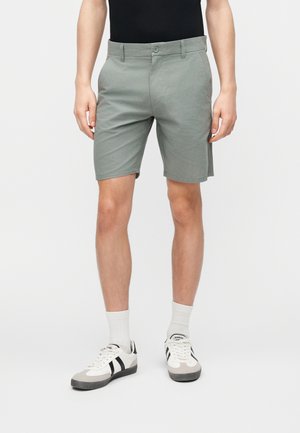 Hombre vestido con pantalones cortos de color gris claro hasta la rodilla, calcetines blancos hasta la pantorrilla y zapatillas blancas con rayas negras, de pie contra un fondo liso.