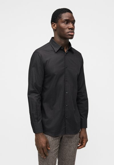 Camisa preta de botões com um padrão texturizado subtil, mangas compridas, design com gola e botões pretos, combinada com calças castanho-claro.