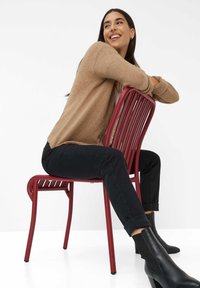 Sedia di metallo rossa con listelli verticali, abbinata a un maglione beige e a jeans neri slim-fit. Presenta eleganti stivaletti neri alla caviglia. Sfondo bianco semplice.