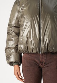 Olivgrüne Pufferjacke aus glänzendem Material, mit gerippten Bündchen und elastischem Saum. Der Reißverschluss verläuft in der Mitte und wird mit braunen strukturierten Hosen kombiniert.