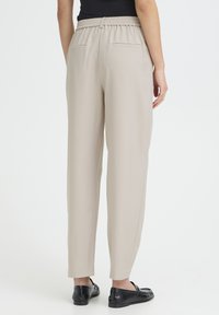 Pantalon beige clair, fuselé, avec une ceinture élastique, des poches arrière et une texture lisse, associé à des mocassins noirs.