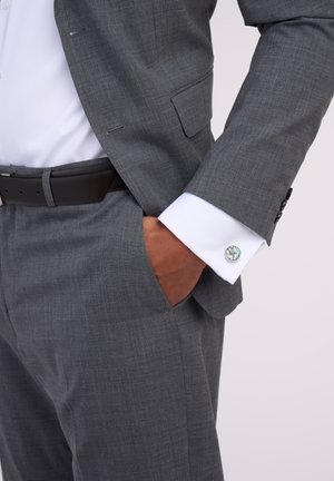 Grauanzug aus strukturiertem Stoff, weißes Hemd und Manschettenknöpfe mit einem runden, bunten Design. Ein schwarzer Gürtel rundet das Outfit ab.