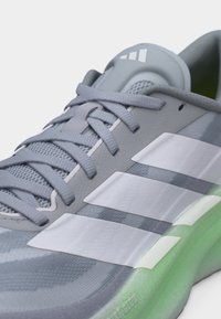 Gros plan d'une chaussure de sport grise avec des bandes blanches, lacets noués, languette rembourrée et semelle amortissante de couleur vert clair.