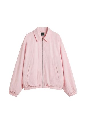 Jeune femme portant une veste rose par-dessus un haut de bikini rose, associée à une jupe blanche et des sandales blanches à talons, debout contre un mur blanc.