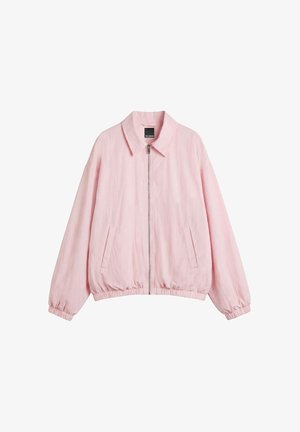 Jeune femme portant une veste rose par-dessus un haut de bikini rose, associée à une jupe blanche et des sandales blanches à talons, debout contre un mur blanc.