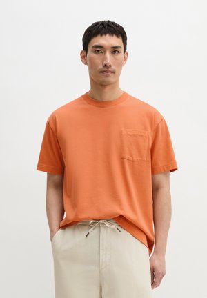 Mann steht, trägt ein oranges kurzärmeliges Hemd mit Tasche und hellbeige Hose mit Kordelzug, vor einem schlichten hellen Hintergrund.