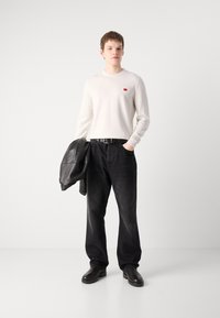 Chemise blanche à manches longues avec un petit logo rouge, associée à un pantalon en denim noir et une veste en cuir noire. Des chaussures noires complètent la tenue.
