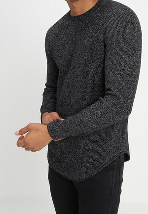 Mann trägt einen dunkelgrauen strukturierten Langarmpullover und schwarze Hose, zieht mit einer Hand den Ärmel zurecht.