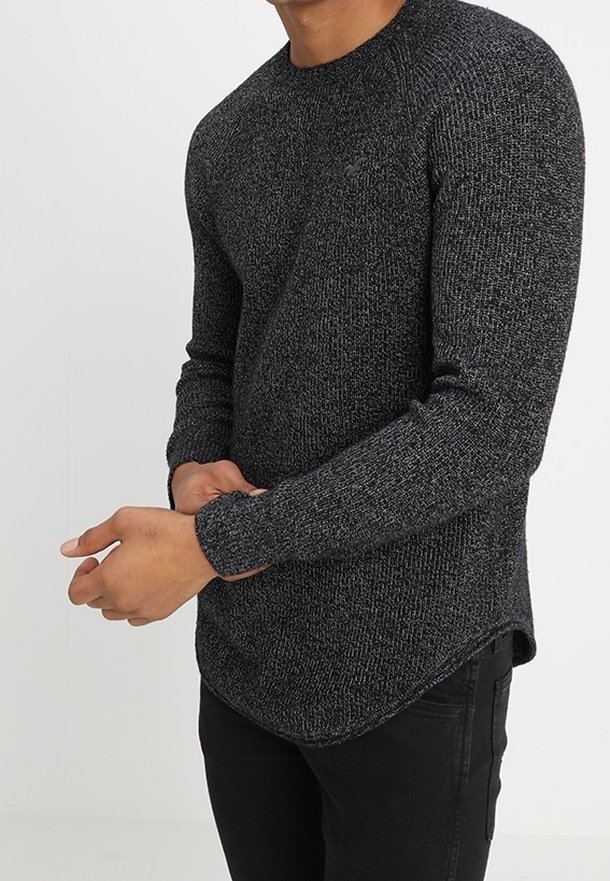 Homme portant un pull à manches longues gris foncé texturé et un pantalon noir, ajustant la manche d'une main.