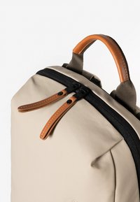 Zaino beige con cerniera nera, inserti in pelle color talpa e tessuto resistente. Presenta una forma sottile e doppi tiranti in pelle per la cerniera.