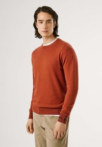 Suéter color óxido con escote redondo, puños y dobladillo acanalados, textura suave. Lleva puesto sobre una camisa blanca, con acentos de logo negro en el puño.