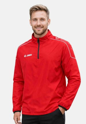 Mann lächelt, trägt eine rote JAKO Sportjacke mit halbem Reißverschluss und schwarze Hosen, steht mit einer Hand in der Tasche vor schlichtem Hintergrund.