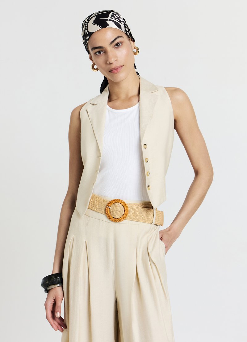 Veste sans manches beige avec un col à revers, t-shirt blanc, pantalon beige plissé, et une large ceinture tissée avec une boucle ronde orange.