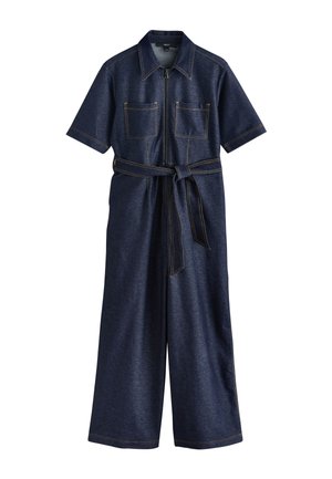 Vue de dos d'une personne portant une robe en denim bleu foncé avec des surpiqûres jaunes et un détail de passants de ceinture à la taille.