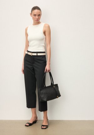 Fehér ujjatlan felsőt, fekete, fehér övvel ellátott, rövidnadrágot és fekete, nyitott orrú magassarkút viselő nő, kezében fekete szőtt táskával, sima fal előtt áll.