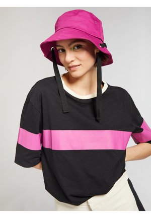 T-shirt nera oversize con una larga striscia orizzontale rosa. Abbinata a un cappello a secchiello rosa brillante con una tracolla nera e dettagli in occhio di bue.