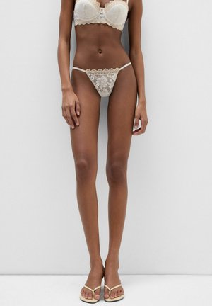 Kobieta w białym koronkowym biustonoszu i pasujących stringach stoi przy białej ścianie, mając na nogach beżowe klapki.