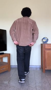 Camisa de couro castanho com riscas verticais, usada sobre calças pretas e ténis. De pé contra uma parede simples com móveis de madeira nas proximidades.