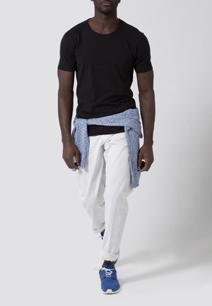 Schwarzes T-Shirt mit kurzen Ärmeln, weiße locker sitzende Hosen, blaue Sportschuhe und ein hellblauer Pullover, um die Taille gebunden.