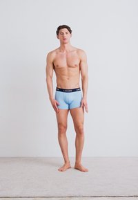 Boxer briefs de algodón azules con un logo negro de Lacoste en la cintura, modelo de pie descalzo contra un fondo blanco liso.