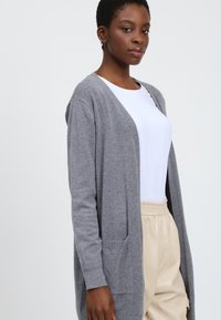Grijze gebreide cardigan met een open voorkant, twee voorzakken en een relaxte pasvorm. Gedragen boven een wit T-shirt en beige broek. Soepele textuur.