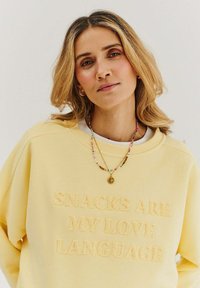 Žltý sveter s mäkkým textom "SNACKS ARE MY LOVE LANGUAGE." Má voľný strih a zaoblený výstrih, kombinovaný s náhrdelníkmi.