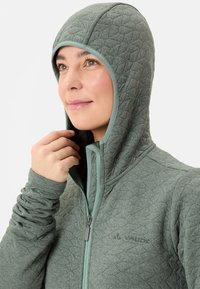 Gequilted groene jas met een capuchon, voorzien van een rits en contrasterende lichtgroene afwerking. De stof heeft een gestructureerd ruitpatroon.