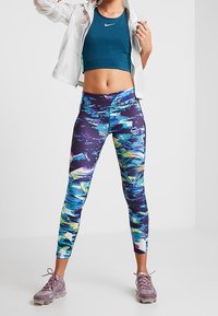 Turkusowy stanik sportowy, biały rozpinany jacket oraz kolorowe legginsy w abstrakcyjny wzór. Szare buty sportowe z sznurówkami. Gładkie tekstury materiałów.