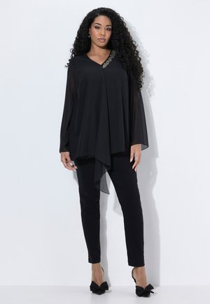 Vrouw met lang, krullend haar draagt een vloeiende zwarte top met gouden versieringen, strakke zwarte broek en zwarte hakken met strikken, staande tegen een witte achtergrond.