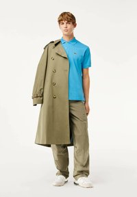 Vihreä trench-coat nappeineen, sininen polo-paita, cargo-housut ja valkoiset lenkkikengät. Yksinkertainen muotoilu ja sileä pinta, paita sisältää logon.