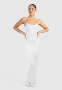 Kvinde står med hænderne på hofterne iført en tætsiddende lysegrå ærmeløs maxikjole og hvide sneakers mod en ensfarvet baggrund.