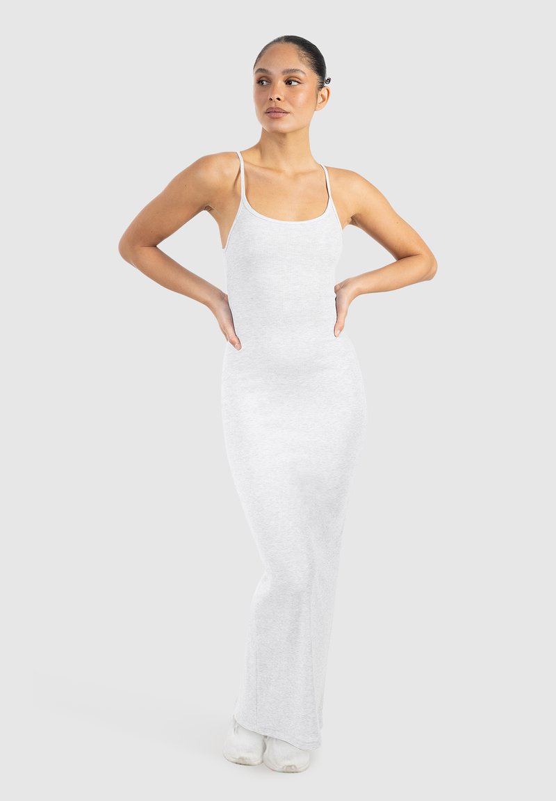 Kvinde står med hænderne på hofterne iført en tætsiddende lysegrå ærmeløs maxikjole og hvide sneakers mod en ensfarvet baggrund.