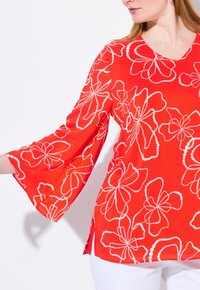 Blouse rouge en tissu léger, avec des motifs floraux blancs. Elle a un col en V, des manches cloche trois-quarts et des fentes latérales.