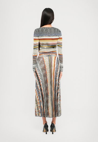Donna con i capelli lunghi e scuri, che indossa un maglione a maniche lunghe a righe multicolori e una gonna midi plissettata coordinata, scarpe con il tacco alto nere, di spalle.