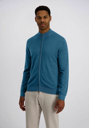 Mann trägt einen blauen Reißverschluss-Pullover und helle beige Hose und steht vor einem schlichten weißen Hintergrund.