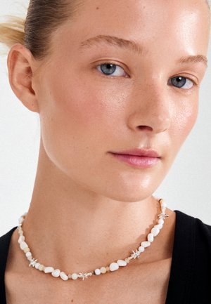 Joven mujer de piel clara y ojos azules que lleva un collar blanco con cuentas y pequeños colgantes en forma de estrella de mar, y una camiseta negra.