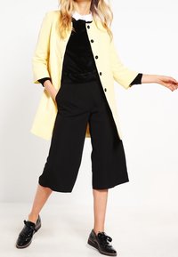Donna che indossa un cappotto giallo pallido, pantaloni neri a gamba larga e taglio corto, top in velluto nero con colletto bianco e scarpe lucide nere con lacci.