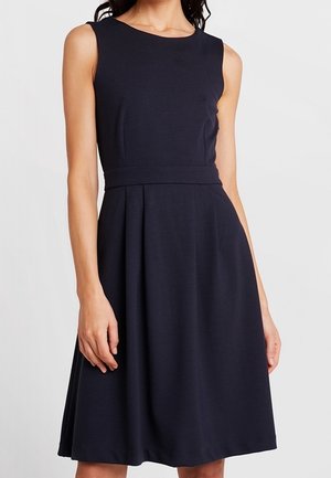 Robe noire sans manches, longueur genou, taille cintrée et légers plis, portée par une femme les bras détendus le long du corps.