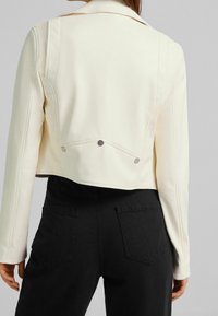 Krótkia kremowa kurtka z usztywnionym kołnierzem, z zaostrzonymi szwami i srebrnymi detalami, zestawiona z czarnymi spodniami z tylnymi kieszeniami.