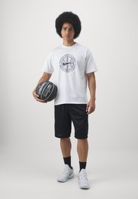 Witte T-shirt met korte mouwen en zwart grafisch logo, gecombineerd met zwarte basketbalsorts. De persoon houdt een basketbal vast en draagt witte sneakers.