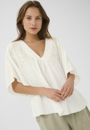 Top alb, ușor, cu decolteu în V și mâneci lejere, având broderie florală discretă în nuanțe de bej. Textură moale, cu un tiv ușor desfăcut.