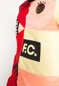 Nike fotbollsdräkt med ett geometriskt mönster i röda, rosa och gula nyanser. Svart "F.C."-text och logotyp med stjärnor som står i förgrunden.