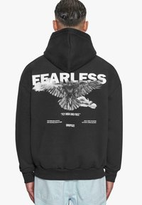 Schwarzer Hoodie mit einem großen weißen Aufdruck eines Adlers und dem Wort "FEARLESS" auf der Rückseite, ergänzt durch weiteren Text und einem gerippten Saum.