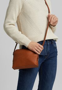 Vrouw in een crèmekleurige gebreide trui en blauwe jeans houdt een bruine leren schoudertas met een lange strap over haar lichaam.