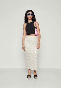 Kvinde iført en sort udenærmet crop top, hvid perforeret midi-skirt, sorte sandaler, blå solbriller og bærende en lille lyserød skuldertaske.