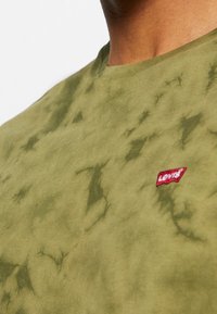 Gros plan d'une personne portant un t-shirt tie-dye vert olive avec un petit patch logo rouge Levi's sur la partie gauche de la poitrine.